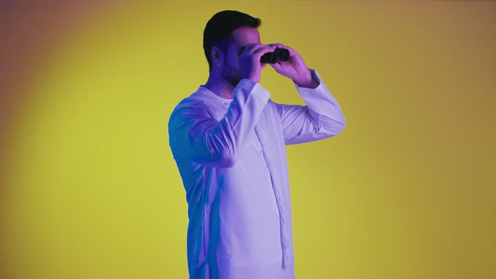 Love of exploration and adventure, gestures of enjoyment and pleasure, seeing distant things through a telescope, a picture of a smiling Emirati Gulf Arab man wearing a kandura holding binoculars, pointing his index finger upwards looking at something, yellow background.