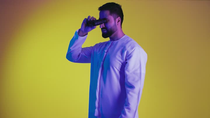 Seeing distant objects through a telescope, gestures of enjoyment and pleasure, a picture of a smiling Emirati Gulf Arab man wearing a kandura holding binoculars, pointing his index finger upwards looking at something, love of exploration and adventure, yellow background.