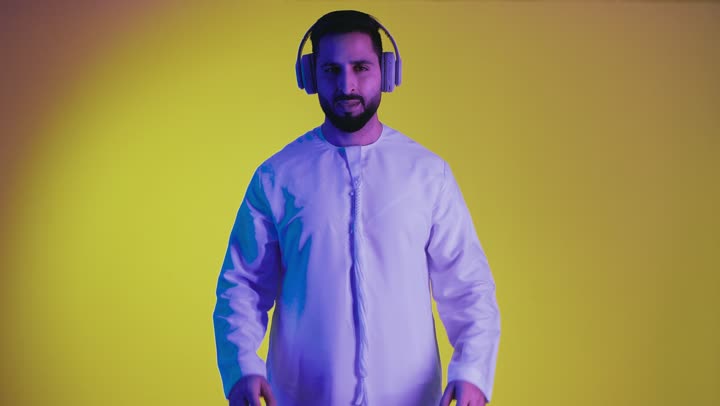Listening to music with expressions of happiness and pleasure, wearing headphones, a picture of an Emirati Gulf Arab man wearing a kandura using headphone technology, having a good time listening to songs and audio clips, taking advantage of modern technology, yellow background.