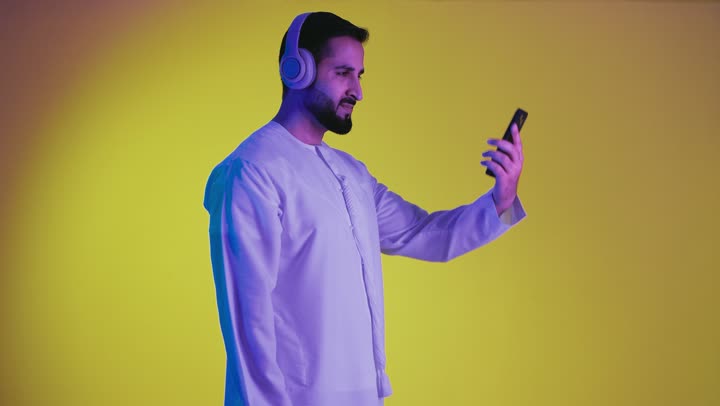 Making video calls, listening to music and audio recordings, using mobile phones and technological devices, a picture of a young Emirati Gulf Arab man wearing a kandura and putting on headphones with happy gestures, using headphone technology, yellow background.