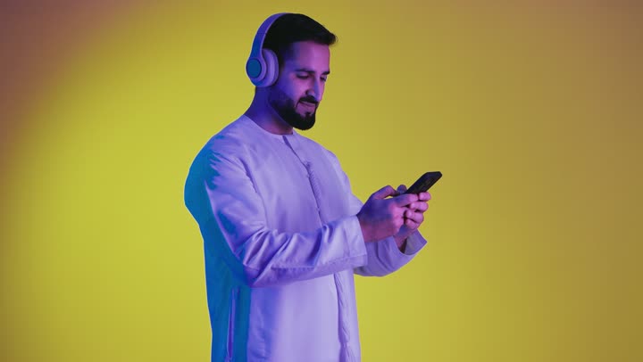 Listening to music and audio recordings, making use of mobile phones and technological devices, a picture of a young Emirati Gulf Arab man wearing a kandura and putting on headphones with happy gestures, using headphone technology, yellow background.