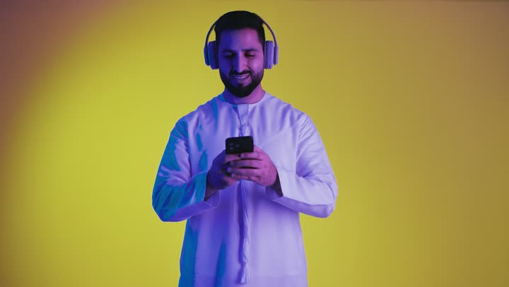 الاستفادة من الجوال والأجهزة التكنولوجية، الاستماع إلى الموسيقى والتسجيلات الصوتية، تصوير لشاب عربي خليجي اماراتى يرتدي الكندورة ويضع سماعة الرأس بإيماءات السعادة, استخدام تقنية الهيدفون، خلفية صفراء .