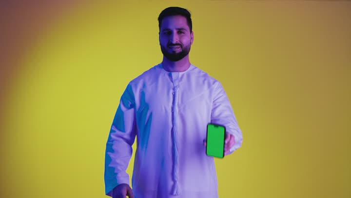 Digital marketing, displaying mobile applications and services, displaying a blank screen on a mobile phone, using advanced technology, photographing a smiling young Emirati Gulf Arab man wearing a kandura looking at the camera, yellow background.
