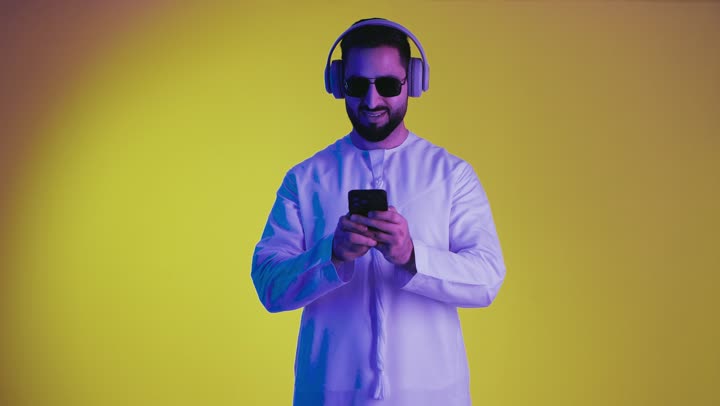 الاستماع إلى الموسيقى والتسجيلات الصوتية، الاستفادة من الجوال والأجهزة التكنولوجية، تصوير لشاب عربي خليجي اماراتى يرتدي الكندورة ويضع سماعة الرأس بإيماءات السعادة, استخدام تقنية الهيدفون، خلفية صفراء .