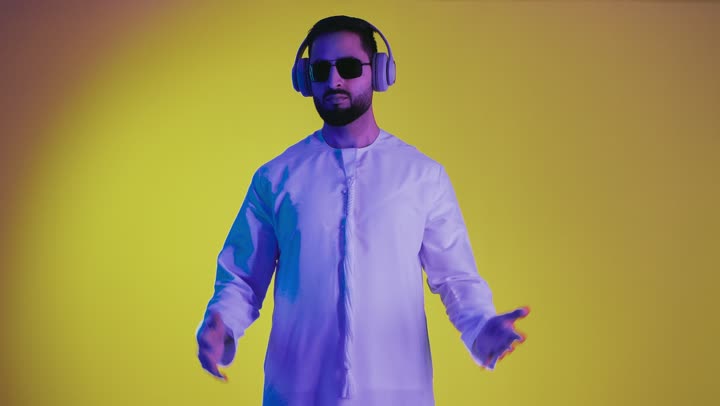 Lifting a specific object, the concept of elegance and attention to one's appearance, the expression of holding and carrying a specific item, a photograph of an Emirati Gulf Arab young man wearing a kandura, black sunglasses, and a headset, opening his hands to display three-dimensional holographic elements, against a yellow background.
