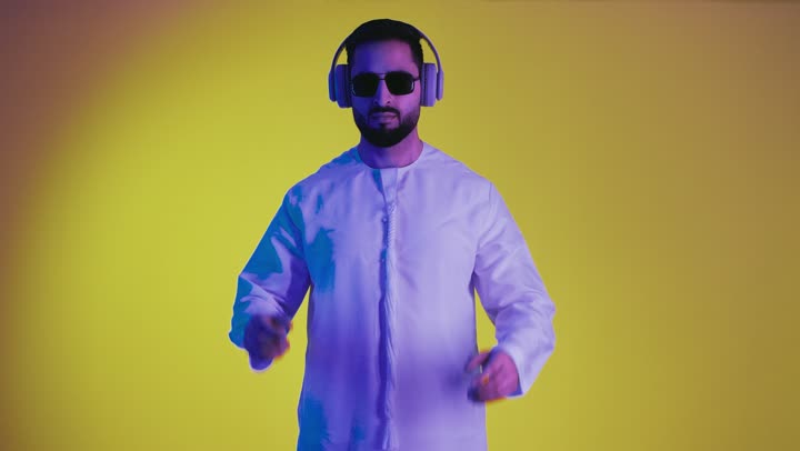رفع شيء معين, مفهوم الاناقة والاهتمام بالمظهر الخارجي, تعابير حمل و إمساك عنصر محدد ، تصوير لشاب عربي خليجي اماراتي يرتدي الكندورة ونظاره سوداء وهيدفون يفتح يديه لعرض عناصر هولوجرامية ثلاثية الأبعاد، خلفية صفراء .