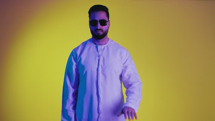 الأمر بالصمت التام، النهي والزجر والانصات، طلب التوقف عن الكلام والصمت، وضع اصبع السبابة أمام الفم بإيماءات الجدية، تصوير لرجل عربي خليجي اماراتي يرتدي الكندورة ونظاره سوداء ينظر للكاميرا بإيماءات الغضب، الجسم بالطول الكامل، خلفية صفراء.