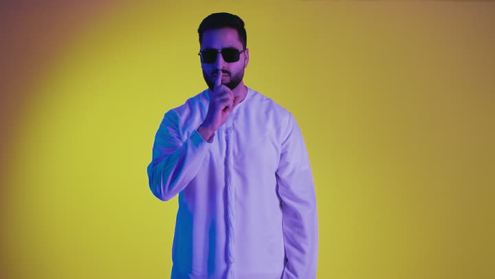 النهي والزجر والانصات، طلب التوقف عن الكلام والصمت، وضع اصبع السبابة أمام الفم بإيماءات الجدية، الأمر بالصمت التام، تصوير لرجل عربي خليجي اماراتي يرتدي الكندورة ونظاره سوداء ينظر للكاميرا بإيماءات الغضب، الجسم بالطول الكامل، خلفية صفراء.