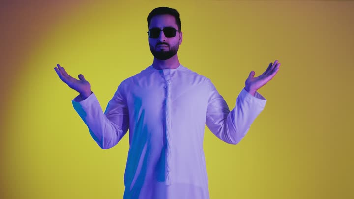 مفهوم الاناقة و الاهتمام بالمظهر الخارجي, رفع شيء معين, تعابير حمل و إمساك عنصر محدد ، تصوير لشاب عربي خليجي اماراتي يرتدي الكندورة ونظاره سوداء يفتح يديه لعرض عناصر هولوجرامية ثلاثية الأبعاد، خلفية صفراء .