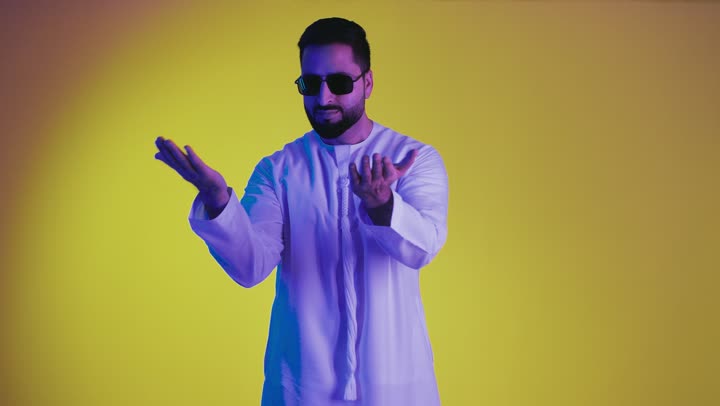 رفع شيء معين, تعابير حمل و إمساك عنصر محدد ، مفهوم الاناقة و الاهتمام بالمظهر الخارجي, تصوير لشاب عربي خليجي اماراتي يرتدي الكندورة ونظاره سوداء يفتح يديه لعرض عناصر هولوجرامية ثلاثية الأبعاد، خلفية صفراء .