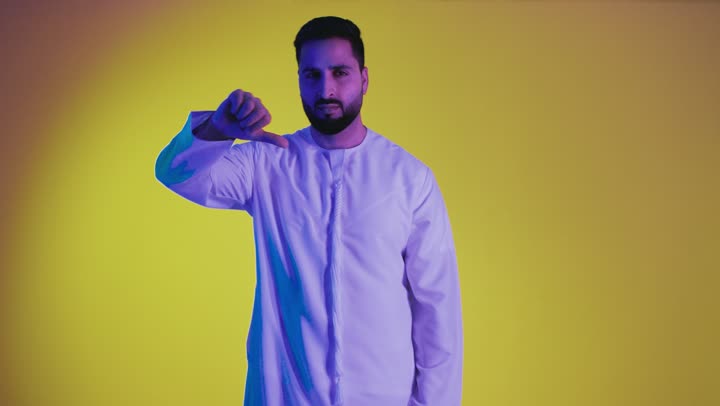 Gestures of dislike and negative evaluation, feeling annoyed by something, hand movements indicating rejection, non-acceptance and failure, a picture of an Arab Gulf Emirati man wearing a kandura pointing his thumb down, yellow background.