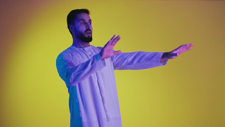 Protecting yourself from external harm, requesting to stop, refrain, and not approach, a picture of an Arab Gulf Emirati man wearing a kandura looking at the camera and raising his hands with expressions of fear and distancing, yellow background.
