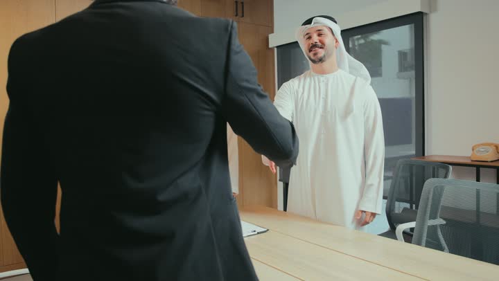 A new business agreement between partners, a successful joint collective deal, presenting ideas and solutions, two Emirati Arab businessmen wearing kanduras and ghutras shaking hands with a young man wearing a formal suit in the meeting room, greetings, greetings and introductions, conducting a job interview and test, exchanging information and experiences