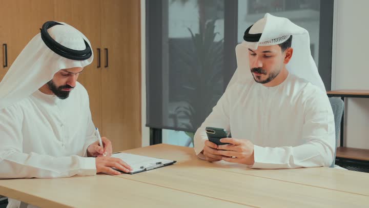 إجراء مقابلة توظيف واختبار, بيئة العمل المريحة في الشركات الإماراتية, رجل يستخدم الهاتف المحمول, تبادل المعلومات والخبرات, اجتماع عمل في غرفة الاجتماعات، رجلا أعمال عربيان خليجيان إماراتيان يرتديان الكندورة والغترة يقومان بتوقيع عقد وصفقة جديدة