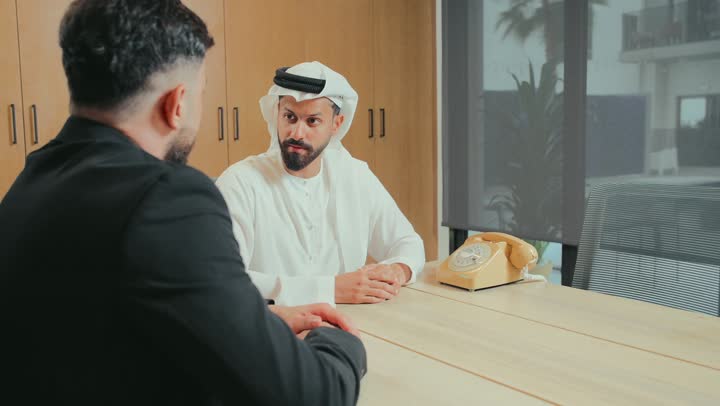 اجراء مقابلة توظيف واختبار، تبادل المعلومات والخبرات، رجل اعمال عربي خليجي إماراتي يرتدي الكندورة والغترة الإماراتية يقابل شاب يرتدي البدلة الرسمية في مقر الشركة، عقد اتفاقية لمشروع جديد، الموافقة على شروط العمل، الرد على مكالمة هاتفية عبر هاتف ثابت سلكي قديم