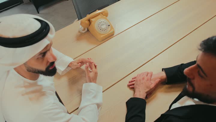 Conducting a job interview and testing, exchanging information and experiences, an Emirati Gulf Arab businessman wearing an Emirati kandura and ghutra meeting a young man wearing a formal suit at the company headquarters, concluding an agreement for a new project, agreeing to the terms of employment, answering a phone call on an old wired landline