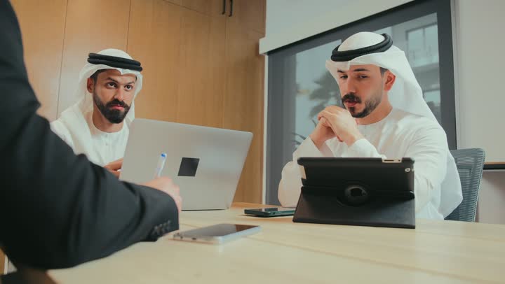 التفاهم والتعاون بين الزملاء لتحقيق أهداف الشركة, بيئة العمل المريحة في الشركات الإماراتية, استخدام الأجهزة التقنية الحديثة في المكاتب, حوار ونقاش لتحقيق الاستراتيجيات والغايات, اجتماع عمل بين موظفين عرب خليجيين اماراتيين في غرفة الاجتماعات، توقيع عقد وصفقة جديدة