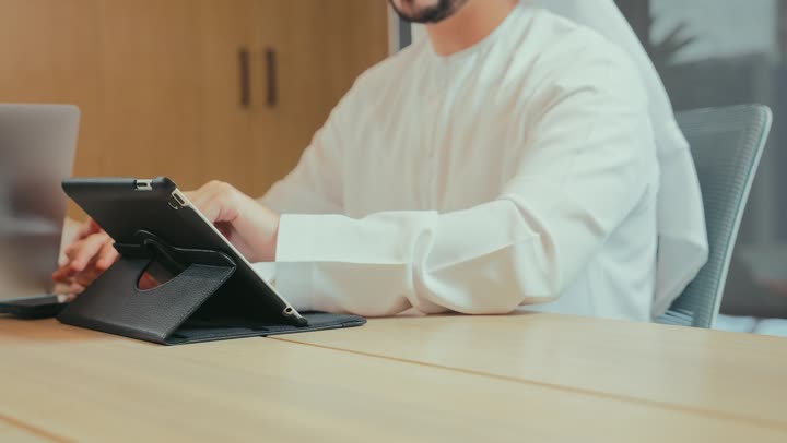 إدارة وإنجاز المهام عن بعد, استخدام الأجهزة والتقنيات الحديثة والمتطورة في المكاتب, تصوير مقرب لرجل عربي خليجي إماراتي يرتدي الكندورة والغترة الإماراتية يعمل على جهاز التابلت اللوحي, مفهوم العمل عبر الأونلاين, مهن ووظائف مكتبية إماراتية