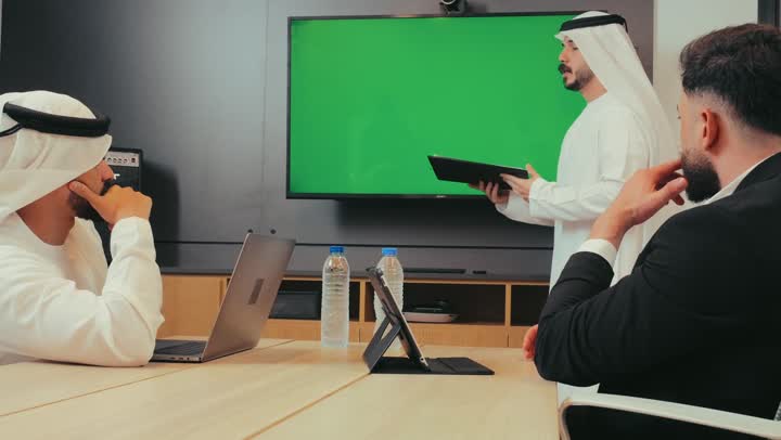 عرض شاشة كروما خضراء فارغة, دمج الأجهزة التقنية الحديثة بالأعمال لسهولة توصيل المعلومة، إدارة و تقسيم المهام، رجل عربي خليجي إماراتي يقوم بشرح المشروع داخل غرفة الاجتماعات للموظفين، فريق عمل يقوم بإجراء اجتماع لتطوير المشاريع و الاستثمارات في الشركة