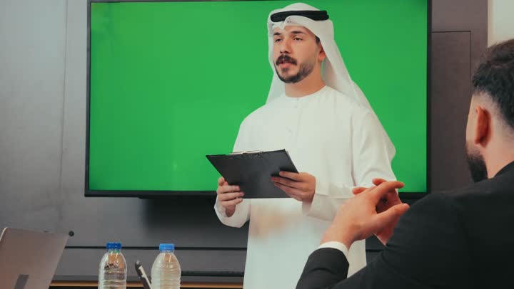 Managing and dividing tasks, an Emirati Gulf man explains the project inside the meeting room for employees, a work team that conducts a meeting to exchange experience and develop projects and investments, display an empty green chroma screen, merge modern technical devices with business to ease the connection of the information