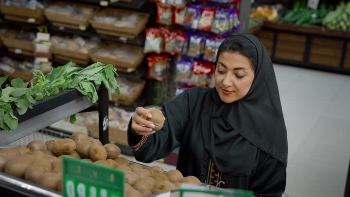 الاستمتاع بقضاء وقت ممتع في التسوق, التجول في إحدى المحلات التجارية في الامارات, تصوير مقرب من الامام لسيدة عربية خليجية اماراتية  مبتسمة محجبة ترتدي العباءة تقوم باختيارالكيوي من قسم الخضار والفواكه, محاصيل زراعية ومنتجات محلية ومستوردة في الماركت