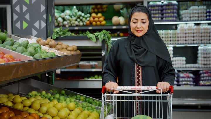 التمتع بالتخفيضات والعروض والخصومات على المنتجات، الاستمتاع بالتجول في المول والمركز التجاري، شراء الاحتياجات و المستلزمات المنزلية للمستهلك، سيدة سعودية تقوم بانتقاء المانجو الطازج، تصوير لامراة عربية خليجية امارتية ترتدي الحجاب والعباءة تجرعربة التسوق أمامها بايماءات السعاده و السرور