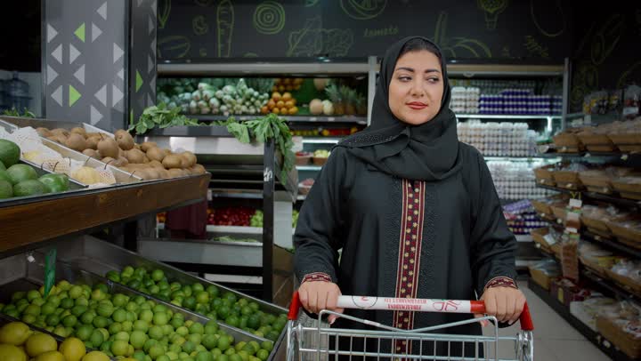 الاستمتاع بالتجول في المول والمركز التجاري، شراء الاحتياجات و المستلزمات المنزلية للمستهلك، سيدة سعودية تقوم بانتقاء المانجو الطازج، تصوير لامراة عربية خليجية امارتية ترتدي الحجاب والعباءة تجرعربة التسوق أمامها بايماءات السعاده و السرور