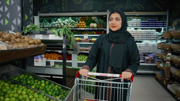 شراء الاحتياجات و المستلزمات المنزلية للمستهلك، الاستمتاع بالتجول في المول والمركز التجاري، سيدة سعودية تقوم بانتقاء التفاح الاخضر الطازج، تصوير مقرب لامراة عربية خليجية امارتية ترتدي الحجاب والعباءة تجرعربة التسوق أمامها بايماءات السعاده و السرور