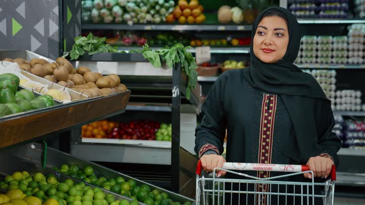 الاستفادة من الخصومات على المنتجات، الاستمتاع بالتجول في المول والمركز التجاري، سيدة سعودية تقوم بانتقاء التفاح الاخضر الطازج، تصوير مقرب لامراة عربية خليجية امارتية ترتدي الحجاب والعباءة تجرعربة التسوق أمامها بايماءات السعاده و السرور