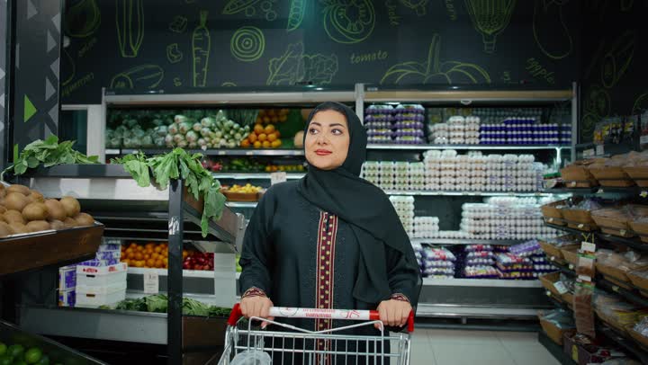 الاستمتاع بالتجول في المول والمركز التجاري، سيدة سعودية تقوم بشراء المستلزمات والحاجيات الضرورية، الاستفادة من الخصومات على المنتجات،  امراة عربية خليجية امارتية ترتدي الحجاب والعباءة تجر عربة التسوق أمامها بايماءات السعاده و السرور