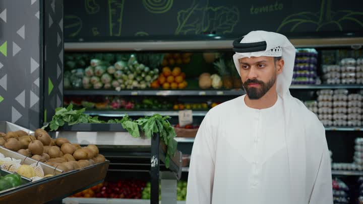 قضاء وقت ممتع في اختيار اللوازم المنزليه, رجل عربي خليجي إماراتي يرتدي الكندورة البيضاء يقوم باختيار التفاح الاخضر أثناء تسوقه داخل أحد السوبرماركت, مأكولات مفيدة صحية على رفوف المتجر، محاصيل زراعيه وطنية اماراتية