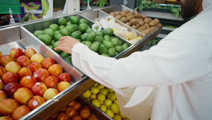 Enjoying shopping in the fruit section of the supermarket, a close-up of the hand of an Emirati Gulf Arab man wearing a white kandura selecting fresh pears, and various types of consumer supplies, including food items and local Emirati agricultural products.