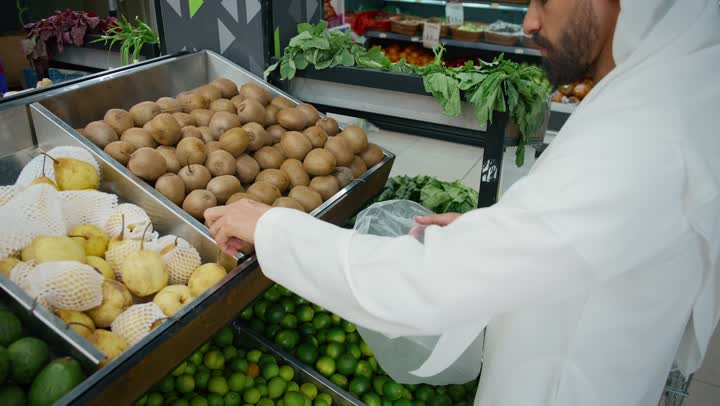 التبضع واختيار الفاكهه الطازجة بعناية من السوبرماركت، تصوير مقرب ليد رجل عربي خليجي إماراتي يرتدي الكندورة البيضاء يقوم بانتقاء الكيوي ، الوعي الغذائي والاهتمام بالتغذية الصحية، منتجات زراعية محليه اماراتية