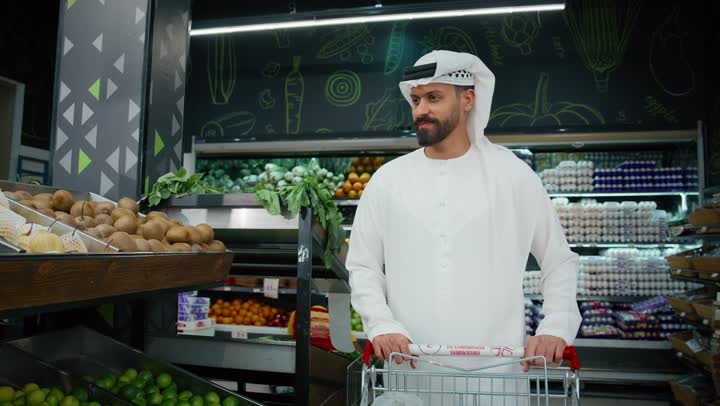 Buying household needs and supplies for the consumer, having a good time in the mall, a close-up of an Emirati Gulf Arab man wearing a white kandura picking green apples while shopping in a supermarket, various goods and food items