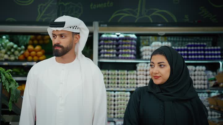 Family cooperation and interest in purchasing fresh food, enjoying shopping with family, an Emirati Gulf Arab couple wearing traditional dress shopping and selecting green apples in the supermarket, Emirati national agricultural crops, various goods and food items