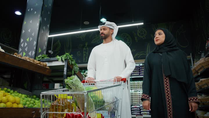 الاختيار الدقيق للخضروات الطازجة، المشاركة في شراء المستلزمات المنزلية، تصويرمقرب لزوجان عربيان خليجيان اماراتيان يرتديان الزي التقليدي يقومان باختيار السلع الغذائية و وضعها في عربة التسوق، محاصيل زراعية وطنيه اماراتية