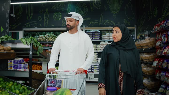 المشاركة في شراء المستلزمات المنزلية، تصويرمقرب لزوجان عربيان خليجيان اماراتيان يرتديان الزي التقليدي يقومان باختيار السلع الغذائية و وضعها في عربة التسوق، الاختيار الدقيق للخضروات الطازجة، محاصيل زراعية وطنيه اماراتية