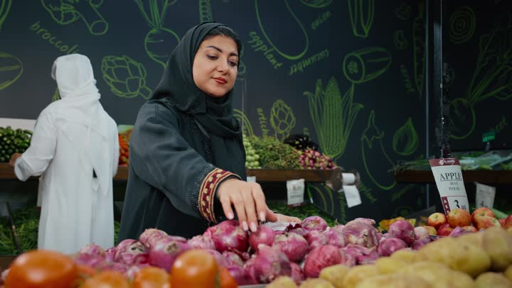 مشاركة الأسرة في توفير وشراء اللوازم والإحتياجات المنزلية، زوجان عربيان خليجيان اماراتيان يرتديان الزي التقليدي يقومان باختيار الخضروات الطازجه في السوبرماركت،تصوير مقرب لامراة عربيه تقوم بانتقاء البصل، محاصيل زراعية وطنيه اماراتية