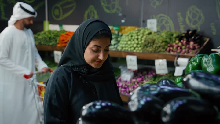 Display of products and food items on shelves in a supermarket, an Arab Emirati Gulf couple shopping, an Arab Gulf Emirati woman wearing a hijab and abaya selecting eggplants, enjoying wandering and buying food items, a local Emirati national agricultural product and crop