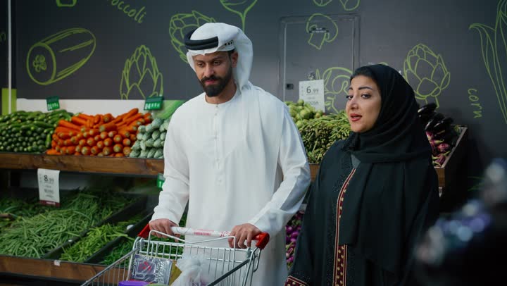 عرض المنتجات والمواد الغذائيه على الارفف في السوبرماركت، زوجان عربيان خليجيان اماراتيان يرتديان الزي التقليدي يقومان بجر عربة التسوق المليئه بالسلع والمستلزمات المنزليه، الاستمتاع في التجول وشراء المواد الغذائية، منتج ومحصول زراعي محلي وطني اماراتي