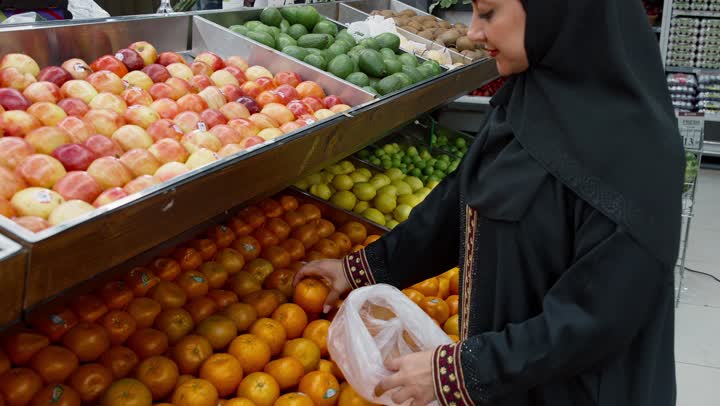 الاهتمام بجودة المنتجات الغذائية أثناء التسوق في السوبرماركت, منتج محلي وطني سعودي, قضاء وقت ممتع في التسوق, شراء الاحتياجات و المستلزمات المنزلية للمستهلك، تصويرمقرب من الجانب لامراة خليجيه اماراتية ترتدي العباءه السوداء تقوم بانتقاء البرتقال، سيده سعوديه تقوم باختيار أجود المنتجات من المركز التجاري