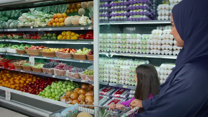 White Friday in UAE stores, the joy of shopping and buying various goods from the supermarket, an Emirati Gulf Arab mother wearing a gray abaya standing with her daughter in front of the fruit shelves, buying household needs, a Saudi woman examining pomegranates before buying, a local national product