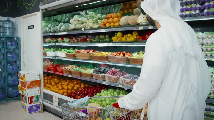 An Emirati Gulf Arab man wearing a kandura and ghutra shopping in the fruits and vegetables section, household needs, goods and food items, choosing good quality fruit, a local Emirati product