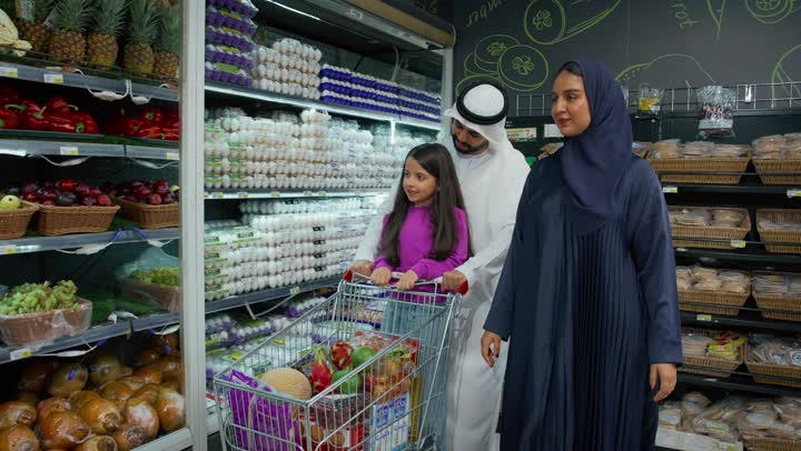 الاستمتاع بالتسوق مع الأبناء، التأثيرالإيجابي للتسوق على العائلة، شراء الإحتياجات المنزلية من السوبرماركت، رجل عربي خليجي اماراتي يرتدي الكندوره والغتره البيضاء برفقة زوجته وابنته، أسرة اماراتيه في قسم الفواكه الطازجة في متجر المواد الغذائية