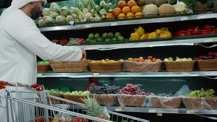 White Friday in UAE stores, the joy of shopping and buying various goods from the supermarket, a Saudi Arabian Gulf father wearing a white kandura and ghutra standing with his daughter in front of the fruit section in one of the UAE stores, shopping cart, buying household needs and consumer supplies
