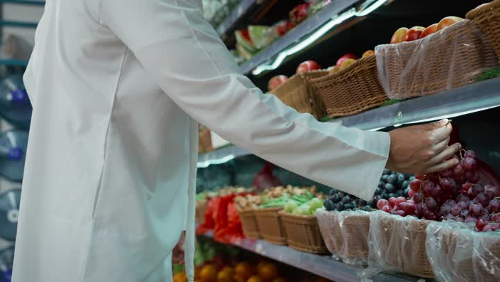 Buying household necessities inside the supermarket, an Emirati man wearing a kandura and ghutra gives his daughter some fruit and puts it in a plastic bag, an Emirati Gulf Arab girl with her father, picking fresh red grapes from the supermarket shelves, local national crops