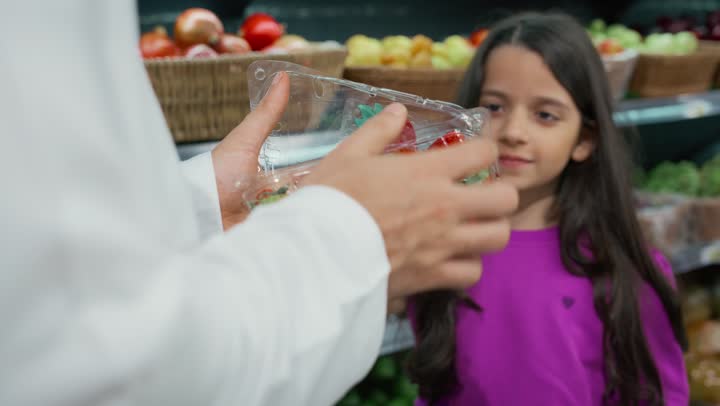 An Emirati Gulf Arab man wearing a white kandura and ghutra gives his daughter fresh strawberries inside the supermarket, a box full of strawberries, taking children while shopping, local national crops