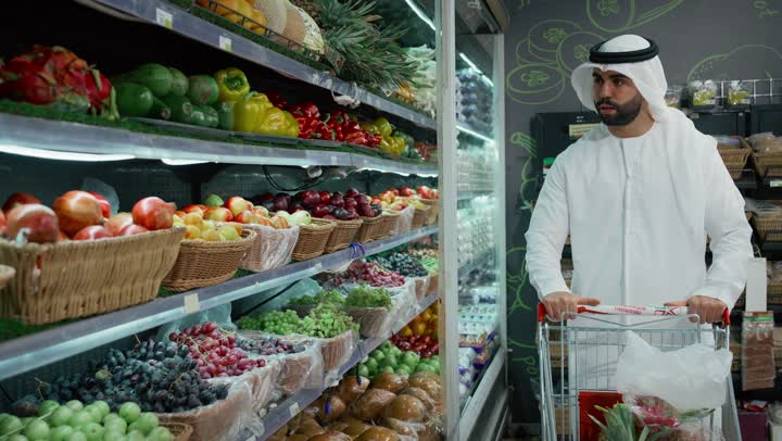 مستلزمات المستهلك من متجر المواد الغذائية، انتقاء أفضل أنواع الفاكهة من السوبرماركت، إيماءات السرور والسعادة بالتسوق، صوره مقربه لرجل عربي خليجي سعودي يرتدي الكندوره والغتره البيضاء يحمل حبة الأفوكادو ويضعها في كيس بلاستيك، منتجات محلية وطنية