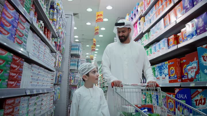 تعليم الأبناء اختيار المواد المناسبة، رجل عربي خليجي إماراتي يرتدي الكندورة والغترة يقوم بالتجول والتبضع في قسم المنظفات في المتجر برفقة ابنه، شراء مستلزمات ومواد التنظيف من سوبرماركت، استغلال العروض والخصومات على المنتجات في المحلات والمتاجر.
