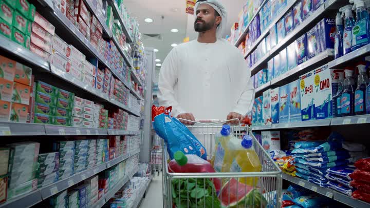 التأكد من شراء السلع المطلوبه في قائمه المستلزمات، امتلاك خيارات اقتناءو استهلاك مختلفة في السوبرماركت، رجل عربي خليجي إماراتي ترتدي الكندورة يقوم يجر عربة التسوق والتجول في قسم المنظفات والمطهرات، قضاء وقت ممتع في اختيار اللوازم المنزلية